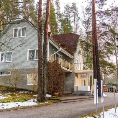 Современный загородный дом с серой фасадной обшивкой, расположенный среди высоких сосен, с заснеженной территорией и аккуратной дорожкой.