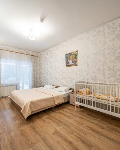 Современная спальня с двуспальной кроватью и детской кроваткой, оформленная в светлых тонах с цветочным узором на стенах. В комнате есть окно с занавесками и деревянный пол, а также зеркало и комод.