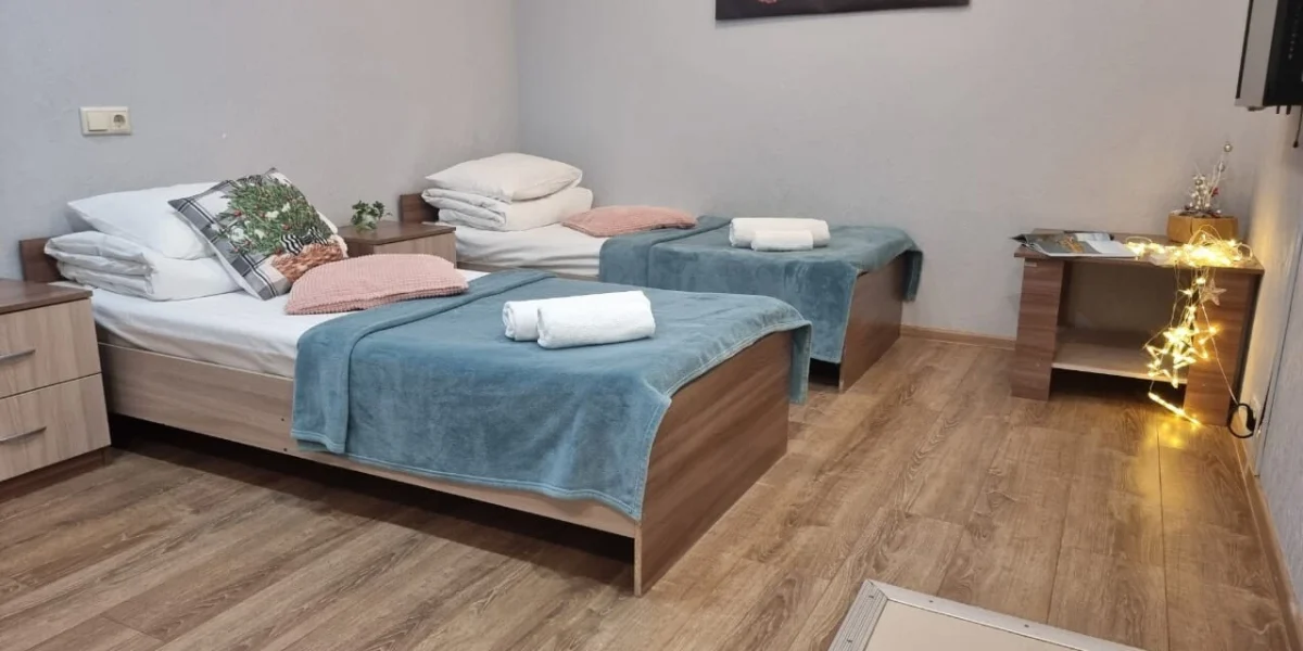 Уютная комната с двумя односпальными кроватями, покрытыми мягкими пледами. На прикроватных тумбочках расположены декоративные подушки и растения. В комнате есть рабочий стол с элементами декора и светящимися огоньками, создающими атмосферу уюта.