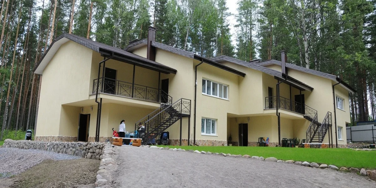 Современный загородный дом с двумя этажами, расположенный в лесу, окруженный зеленью. На переднем плане виден проход и каменная дорожка, а на заднем плане - люди, занимающиеся делами на улице.