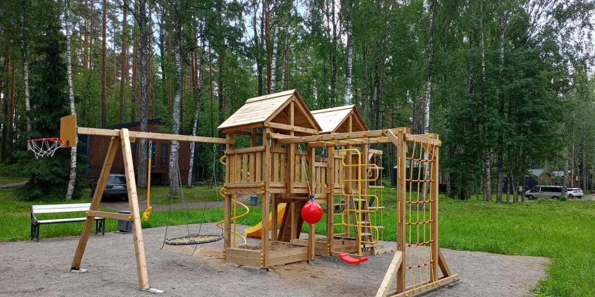 Детская площадка из дерева с горкой, качелями и спортивной корзиной, окруженная зелеными деревьями в парке.